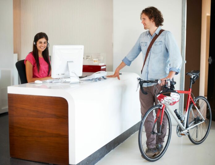 Office, desk and man with bike for business, travel and talking about commute to work with receptionist in morning. Businessman, bicycle and conversation with woman in administration or secretary